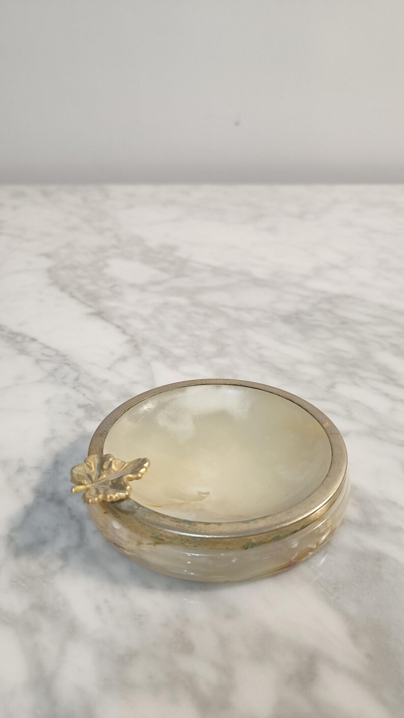 Puede incluir: Un peque&ntilde;o cenicero redondo de alabastro blanco con un dise&ntilde;o de hoja de oro. El cenicero es redondo y tiene un borde ligeramente elevado.