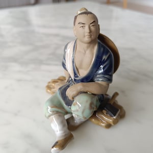 May include: A ceramic figurine of a seated man, wearing a blue and white top and green trousers. The figure has a neutral skin tone and is resting on a wooden base with a hat behind him. The figurine is a decorative item.