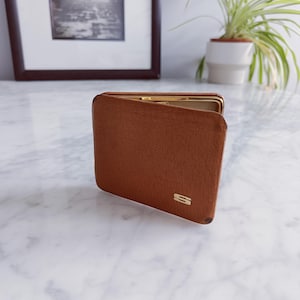May include: A brown wallet with a gold-colored clasp and a small gold-colored "S" emblem. The wallet is made of a textured material and is open, revealing a gold interior. The wallet is sitting on a white marble surface.