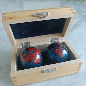 Peut inclure: Une paire de boules métalliques sphériques avec un motif de dragon bleu et rouge, placées dans une boîte rectangulaire en bois clair. La boîte est dotée d'un fermoir et de charnières en métal argenté, avec un intérieur doublé de velours noir.
