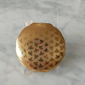 Peut inclure: Un poudrier rond doré avec un motif de papillons. Le poudrier est présenté sur un présentoir en plastique transparent. La surface du poudrier a un aspect texturé.