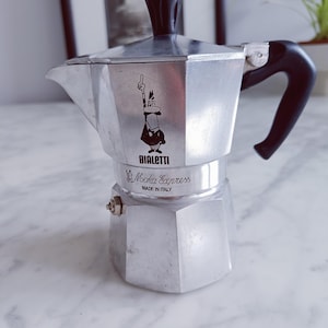 May include: A white ceramic mug with a black handle and the text "But First, Coffee" in a bold, black font. The mug is filled with a dark liquid, probably coffee, and rests on a light-coloured surface.