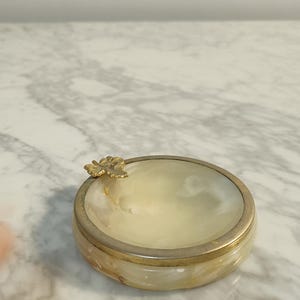 Puede incluir: Un cenicero peque&ntilde;o, redondo, de alabastro blanco con un borde dorado y un dise&ntilde;o de hoja de oro en el lateral.