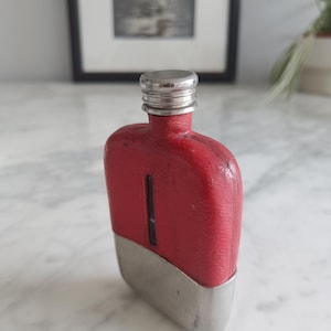Può includere: Una fiaschetta da tasca con un rivestimento rosso testurizzato sulla parte superiore e una base in metallo argentato liscio. La fiaschetta presenta un tappo a vite argentato e una finestrella verticale trasparente al centro della sezione rossa.