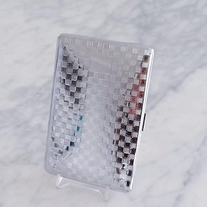 May include: A rectangular, silver-colored cigarette case with a textured, checkered pattern. The case is displayed on a clear plastic stand, showcasing its reflective surface. The case has a small black clasp.