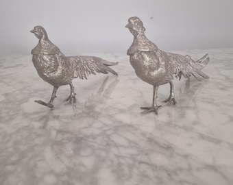 May include: Two silver pheasant figurines standing on a white marble surface. The pheasants are facing different directions and have detailed feathers.