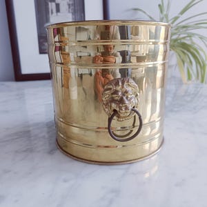 Puede incluir: Un cubo cilíndrico de latón con un asa de anillo con cabeza de león. El cubo tiene un acabado pulido y bandas horizontales. El fondo incluye una imagen en blanco y negro enmarcada y una planta.