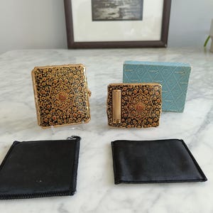May include: A vintage vanity set comprising a compact, lipstick holder, and two black satin pouches. The compact and lipstick holder display a black background with a gold and orange floral pattern. A light blue box is also visible.
