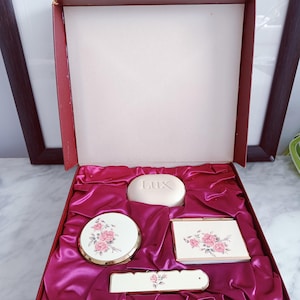 May include: A vintage vanity set presented in a red box with a pink satin lining. The set includes a round compact, a rectangular mirror, a soap bar with "Lux" text, and a nail file, all decorated with pink rose floral patterns.