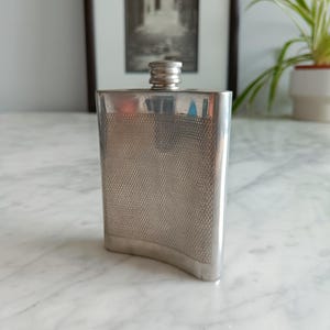 Puede incluir: Una petaca plateada con una superficie texturizada y estampada. La petaca tiene una base curva y un tapón de rosca. El fondo incluye una imagen en blanco y negro enmarcada y una planta en maceta, lo que sugiere un tema vintage o antiguo.