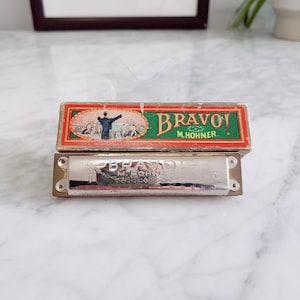 Puede incluir: Una armónica M. Hohner Bravo! vintage con su caja original. La caja es roja, verde y dorada con la palabra "Bravo!" y una ilustración de un director de orquesta. La armónica es plateada.