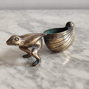 May include: A bronze frog figurine holding a small, shell-shaped dish. The frog is standing on a white marble surface.