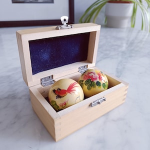 Puede incluir: Una caja de madera clara con tapa con bisagras, que contiene dos esferas decorativas. Una esfera presenta un diseño de pájaro rojo, la otra tiene un estampado floral. La caja tiene un forro de terciopelo azul oscuro.