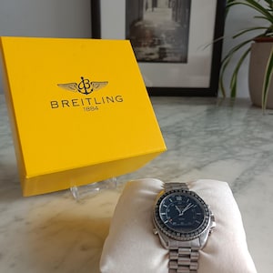 May include: A silver Breitling 1884 wristwatch with a dark dial and metal link band, displayed on a white cushion next to a bright yellow square box featuring the black Breitling logo and the text Breitling 1884 printed on the front surface.
