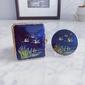 May include: A vintage, rectangular, navy blue enamel case and a matching round compact. Both feature a gold trim and a painted scene of flying birds over a lake with reeds. The case has a hinged lid.