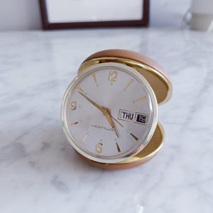 Op de afbeelding: Een vintage Westclox reisklok met een zilveren wijzerplaat, gouden cijfers en wijzers. De klok geeft de dag en datum weer. Het is gevat in een bruine, lederachtige behuizing met een goudkleurige rand. De klok is open.