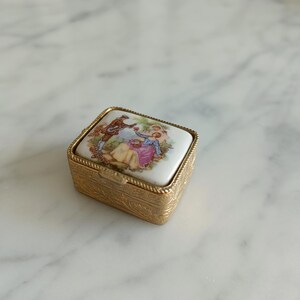 Puede incluir: Un pequeño joyero rectangular con una base de metal dorado que presenta un intrincado patrón de hojas en relieve. La tapa está rematada con una pieza de cerámica blanca que muestra una colorida ilustración de dos figuras en un jardín.
