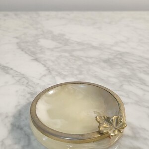 Puede incluir: Un cenicero de alabastro blanco con un dise&ntilde;o de hoja de oro. El cenicero tiene un borde plateado.