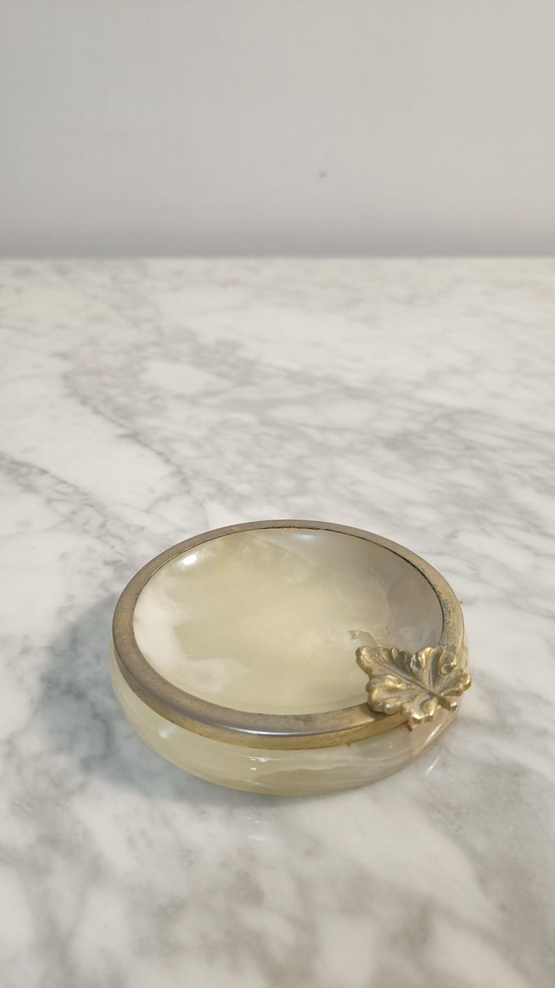 Puede incluir: Un cenicero de alabastro blanco con un dise&ntilde;o de hoja de oro. El cenicero tiene un borde plateado.