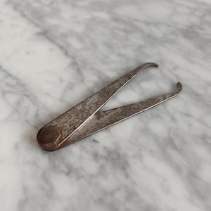 May include: A vintage, rusty metal caliper tool with curved tips. The tool has a hexagonal pivot and is designed for measuring. The metal has a weathered appearance, suggesting age and use. The tool is lying on a marble surface.