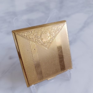 May include: A gold-colored, square compact with intricate engraved patterns. The compact features a central floral design and vertical and diamond-shaped textures. It is displayed on a clear stand, showcasing its ornate details.