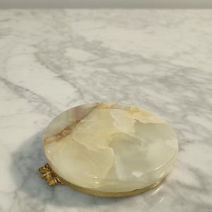Puede incluir: Una caja de baratijas redonda de m&aacute;rmol blanco y marr&oacute;n con un borde y una bisagra de metal dorado.