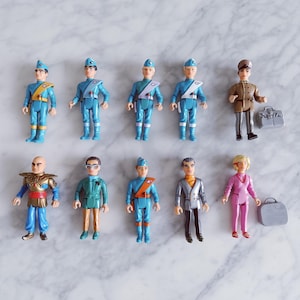 Puede incluir: Una colección de figuras de acción vintage, con varios personajes con uniformes y atuendos detallados. Las figuras son principalmente azules, marrones y rosas, con accesorios como maletines. Los juguetes están dispuestos sobre una superficie de mármol blanco.