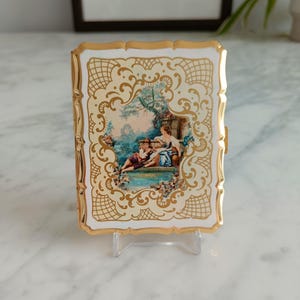 May include: A rectangular, vintage cigarette case with a gold-colored frame and a cream-colored background. The case features a painted scene of two people near a body of water. The edges are decorated with a gold filigree pattern.