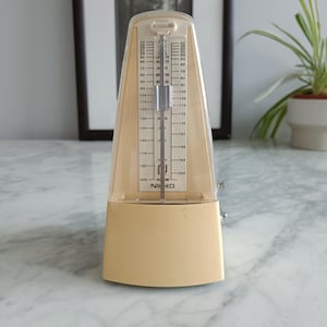 May include: A beige Nikko metronome with a clear plastic cover. The metronome has a scale with numbers from 40 to 208 and a swinging pendulum. The metronome is on a marble surface.