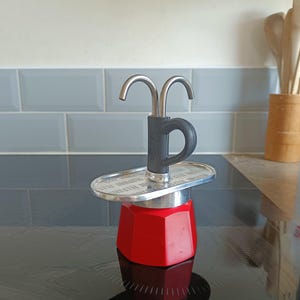 May include: A red octagonal stovetop espresso maker with a silver metallic top plate featuring embossed text. The device includes a grey handle and a dual-pronged silver metal spout extending upwards from the center of the lid assembly.