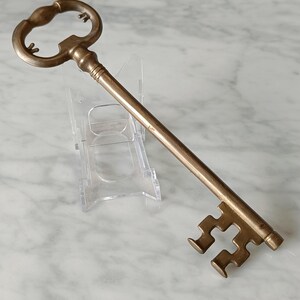 May include: A decorative brass key, featuring an elaborate handle and a detailed key bit. The key is presented on a clear acrylic stand, set against a marble surface. The design suggests a vintage or collectible item.