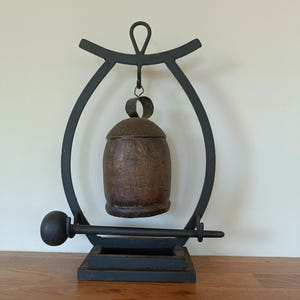 May include: A dark brown, textured metal bell hangs from a black metal arched stand. The stand features a rectangular base and a horizontal bar with a spherical end, positioned to strike the bell. The entire structure is made of dark metal.