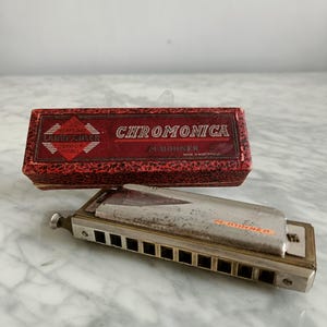 May include: A vintage red and gold harmonica in its original box. The box is labelled "Chromonica" and "M. Hohner" with the text "Junior Model" and "Made in Germany".