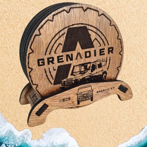 Puede incluir: Posavasos de madera con borde negro y acabado en madera de color marrón. Los posavasos presentan un logotipo en blanco y negro con el texto "GRENADIER" y una imagen en blanco y negro de un coche. Los posavasos están apilados en un soporte de madera con el texto "INEOS GRENADIER" en la parte delantera.