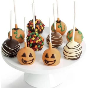 May include: A white cake stand displays a variety of Halloween-themed cake pops. Some are orange with jack-o'-lantern faces, others are decorated with chocolate and sprinkles. Each cake pop is on a white stick.