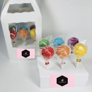 May include: A white box with a window showing six colorful cake pops. The cake pops are on sticks and are decorated with white sprinkles. The box has a pink label with the text "The Cake Pop Co."
