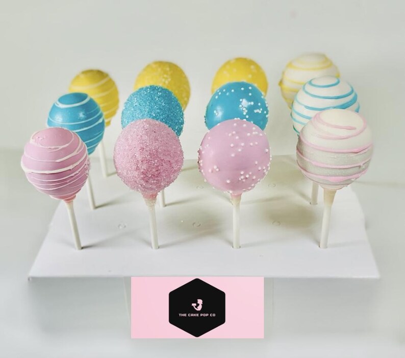 Peut inclure: Un pr&eacute;sentoir &agrave; cake pops blanc avec 12 cake pops d&eacute;cor&eacute;s en rose, bleu et jaune. Les cake pops sont dispos&eacute;s en cercle sur le pr&eacute;sentoir. Le pr&eacute;sentoir porte un logo rose et noir qui dit "The Cake Pop Co."