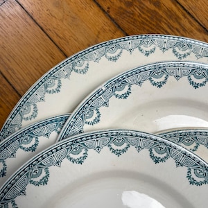 Peut inclure: Plusieurs assiettes en céramique blanche avec une bordure décorative turquoise et blanche. Les assiettes sont disposées sur une surface en bois, mettant en valeur leur design détaillé. La bordure turquoise présente un motif répétitif de formes géométriques et d'éléments floraux.