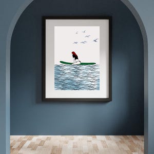 May include: Framed art print featuring a minimalist illustration of a person on a green surfboard in blue water. The artwork includes stylized waves, a red-haired figure, and several birds in the sky. The print is displayed on a blue wall.