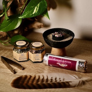 Può includere: Un assortimento di oggetti per pratiche spirituali, tra cui due piccoli barattoli di vetro con coperchi dorati, una ciotola nera su un supporto di legno, una confezione di tavolette di carbone, una piuma e delle pinze metalliche. Piante verdi e frondose sullo sfondo.