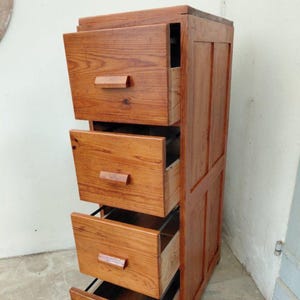 Può includere: Un mobile alto in legno con quattro cassetti, ciascuno con una maniglia rettangolare in legno. Il mobile è realizzato in legno marrone chiaro, con venature visibili. Tre cassetti sono aperti, rivelando l'interno. Il mobile è progettato per lo stoccaggio.