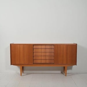 Puede incluir: Aparador de madera de estilo moderno de mediados de siglo con acabado marrón claro. El mueble tiene dos puertas correderas y un conjunto de cinco cajones en el centro. El aparador está sostenido por cuatro patas cónicas.