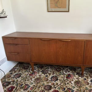 Könnte beinhalten: Ein Sideboard aus Holz im Mid-Century-Stil mit drei Schubladen links und zwei Schranktüren rechts. Das Holz hat einen warmen, rötlich-braunen Farbton. Das Sideboard steht auf angewinkelten Beinen.