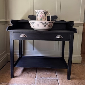 May include: A black washstand with a drawer and a lower shelf. A floral-patterned pitcher and bowl sit on top. The pitcher and bowl have pink and green floral designs with black trim. The washstand has a decorative back.