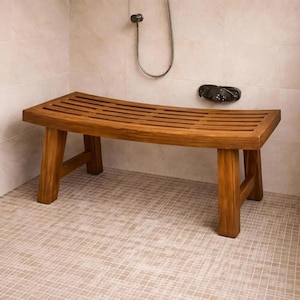 May include: A wooden shower bench with a curved, slatted seat and four sturdy legs. The bench is made of light brown wood and is placed in a tiled shower stall. The showerhead and soap dish are visible in the background.