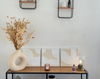 May include: Three beige and white abstract wall art pieces with a raised, textured design. The art is displayed on a shelf with a vase of dried pampas grass, a candle, and a lamp.