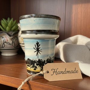 Puede incluir: Una maceta de cerámica hecha a mano con un diseño de paisaje. La maceta presenta un cielo azul, una silueta de árbol negro y una base amarilla. Una etiqueta con la palabra "Handmade" está adjunta.