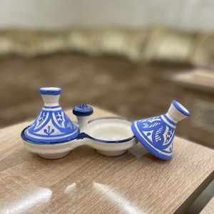 May include: A ceramic serving dish with a blue and white design. The dish has a central section with two small, lidded containers on either side. The lids and containers are decorated with a blue and white pattern. The dish is on a wooden surface.