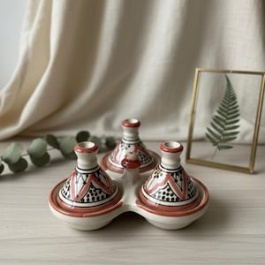 Puede incluir: Una fuente de cerámica blanca con tres pequeños recipientes de tagine con tapa. Los recipientes tienen diseños geométricos rojos, negros y blancos. La fuente está sobre una superficie de madera clara.