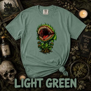 May include: A sage green short-sleeve t-shirt featuring a graphic print of a carnivorous plant with a wide-open mouth, sharp teeth, and a red tongue. The shirt has a Comfort Colors brand label at the inner neckline indicating a size medium.